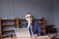 Portrait of grey haired pensive businessman in glasses and blue suit sitting and looking on laptop