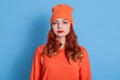 Portrait of grumpy moody girl in hat and warm jumper looking at camera over blue background, female with curls and red