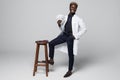 Portrait of handsome Afro American doctor in white coat looking at camera on chair holding dollars, on graybackground Royalty Free Stock Photo