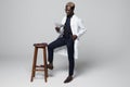 Portrait of handsome Afro American doctor in white coat looking at camera on chair holding dollars, on graybackground Royalty Free Stock Photo