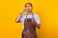 Portrait of handsome asian young man wearing apron holding game controller and thinking about something isolated on yellow Royalty Free Stock Photo