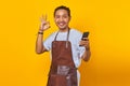 Portrait of handsome asian young man wearing apron holding smartphone and showing ok sign over yellow background