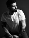 Portrait of handsome bearded brutal muscular man with tattoo on hands in white t-shirt sitting and looking aside Royalty Free Stock Photo