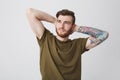 Portrait of handsome bearded caucasian man with tattoo on arm and stylish hairstyle holding hands behind head, looking