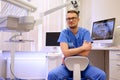 Portrait of a handsome bearded dentist male in glasses wearing a blue uniform, sitting in a dentist office.