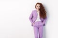 Portrait of handsome beautiful curly redhead with makeup in pink suit standing with hands on pockets and looking at camera with