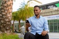 Portrait of handsome black African businessman outdoors in city during summer sitting and using mobile phone while Royalty Free Stock Photo