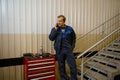 Portrait of a handsome Caucasian man, technician, garage mechanic talking on mobile phone standing near a box with work tools for