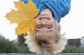 Portrait of a handsome funny blond boy turned upside down, holds a large yellow maple leaf in his mouth