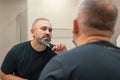 Portrait of handsome gray-haired man in his 40s trimming his beard himself standing in bathroom