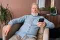 Portrait of handsome gray-haired mature male holding smartphone using mobile online app, looking to screen sitting on Royalty Free Stock Photo