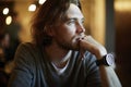 Portrait of handsome hipster guy with long hair and beard sitting in sunny cafe, resting near window. Romantic man looks lonely.