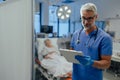 Portrait of handsome male doctor, patient in hospital bed behind. ER doctor examining senior patient, reading her Royalty Free Stock Photo