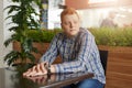 A portrait of a handsome man with red hair wearing checked shirt and jeans sitting at wooden table near green palm holding his han Royalty Free Stock Photo