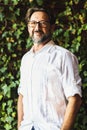 Portrait of handsome mature bearded man with eyeglasses smiling while looking at camera in front of green leaves background. Royalty Free Stock Photo