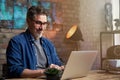 Portrait of handsome mature man with grey hair, wearing red glasses, working on laptop, sitting at desktop at home or in the Royalty Free Stock Photo