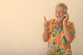 Studio shot of thoughtful happy senior bearded tourist man smiling and laughing while talking on mobile phone and Royalty Free Stock Photo