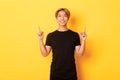 Portrait of handsome smiling asian guy in black t-shirt pointing fingers up and looking satisfied, yellow background