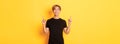 Portrait of handsome smiling asian guy in black t-shirt pointing fingers up and looking satisfied, yellow background