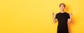 Portrait of handsome smiling asian guy in black t-shirt pointing fingers up and looking satisfied, yellow background