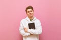 Portrait of handsome student boy with blond hair and in white hoodie stands on pink background and poses at camera with books in Royalty Free Stock Photo