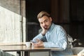 Portrait of handsome thoughtful bearded young man in blue denim shirt sitting in cafe and drinking coffee, tired for waiting Royalty Free Stock Photo