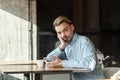 Portrait of handsome thoughtful bearded young man in blue denim shirt sitting in cafe and drinking coffee, tired for waiting