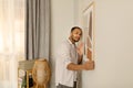 Portrait of handsome young African American man hanging picture frame on white wall at home, decorating his apartment Royalty Free Stock Photo