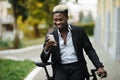 Portrait of handsome young Afro American man in casual clothes using phone, looking away and smiling while leaning on his bike, Royalty Free Stock Photo