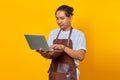 Portrait of handsome young barista using laptop looking serious doing office work on yellow background