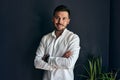 Portrait of handsome young bearded man in stylish casual outfit, posing with crosed arms against black wall in the office. Hipster