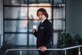 Portrait of handsome young businessman in black suit and tie stands indoors with cup of drink Royalty Free Stock Photo