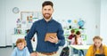 Portrait of handsome young joyful Caucasian male teacher standing in classroom typing on tablet, looking at camera and Royalty Free Stock Photo