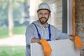 Portrait of handsome young male builder in hard hat looking positive, holding drywall while working at construction site