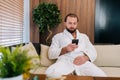 Portrait of handsome young man with beard wearing white bathrobe using mobile phone looking to device screen, sitting by Royalty Free Stock Photo