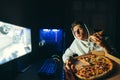 Portrait of handsome young man in hoodie sitting at computer at home at night, watching game streams and eating pizza from Royalty Free Stock Photo