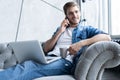 Portrait of handsome young man making a call and using his laptop while sitting on sofa at home Royalty Free Stock Photo