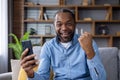 Portrait of a happy African American man sitting on the couch at home, using a mobile phone, and looking happy at the Royalty Free Stock Photo