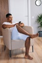 Portrait of happy African-American man sitting at armchair in domestic room and using mobile phone. Royalty Free Stock Photo