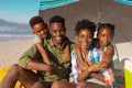 Portrait of happy african american young parents with boy and girl sitting under umbrella at beach Royalty Free Stock Photo