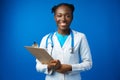 Portrait of happy african medical intern woman doctor with clipboard in blue studio
