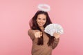 Portrait of happy angelic young woman with halo above head holding dollar banknotes and pointing to camera