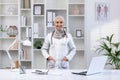 Portrait of happy arab female doctor in hijab standing at table in hospital office with stethoscope around neck, hands Royalty Free Stock Photo