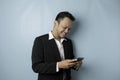 A portrait of a happy Asian businessman is smiling and holding his smartphone wearing black suit isolated by a blue background Royalty Free Stock Photo