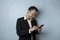 A portrait of a happy Asian businessman is smiling and holding his smartphone wearing black suit isolated by a blue background Royalty Free Stock Photo