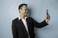 A portrait of a happy Asian businessman is smiling and holding his smartphone wearing black suit isolated by a blue background Royalty Free Stock Photo