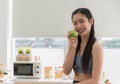 Portrait of happy Asian healthy woman in sportswear holding and eating green apple in kitchen at apartment