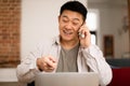 Portrait of happy asian man working from home, talking on cellphone and using laptop, having phone conversation Royalty Free Stock Photo