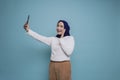 A portrait of a happy Asian Muslim woman wearing a white shirt and hijab, holding her phone, isolated by blue background Royalty Free Stock Photo