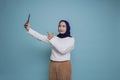 A portrait of a happy Asian Muslim woman wearing a white shirt and hijab, holding her phone, isolated by blue background Royalty Free Stock Photo